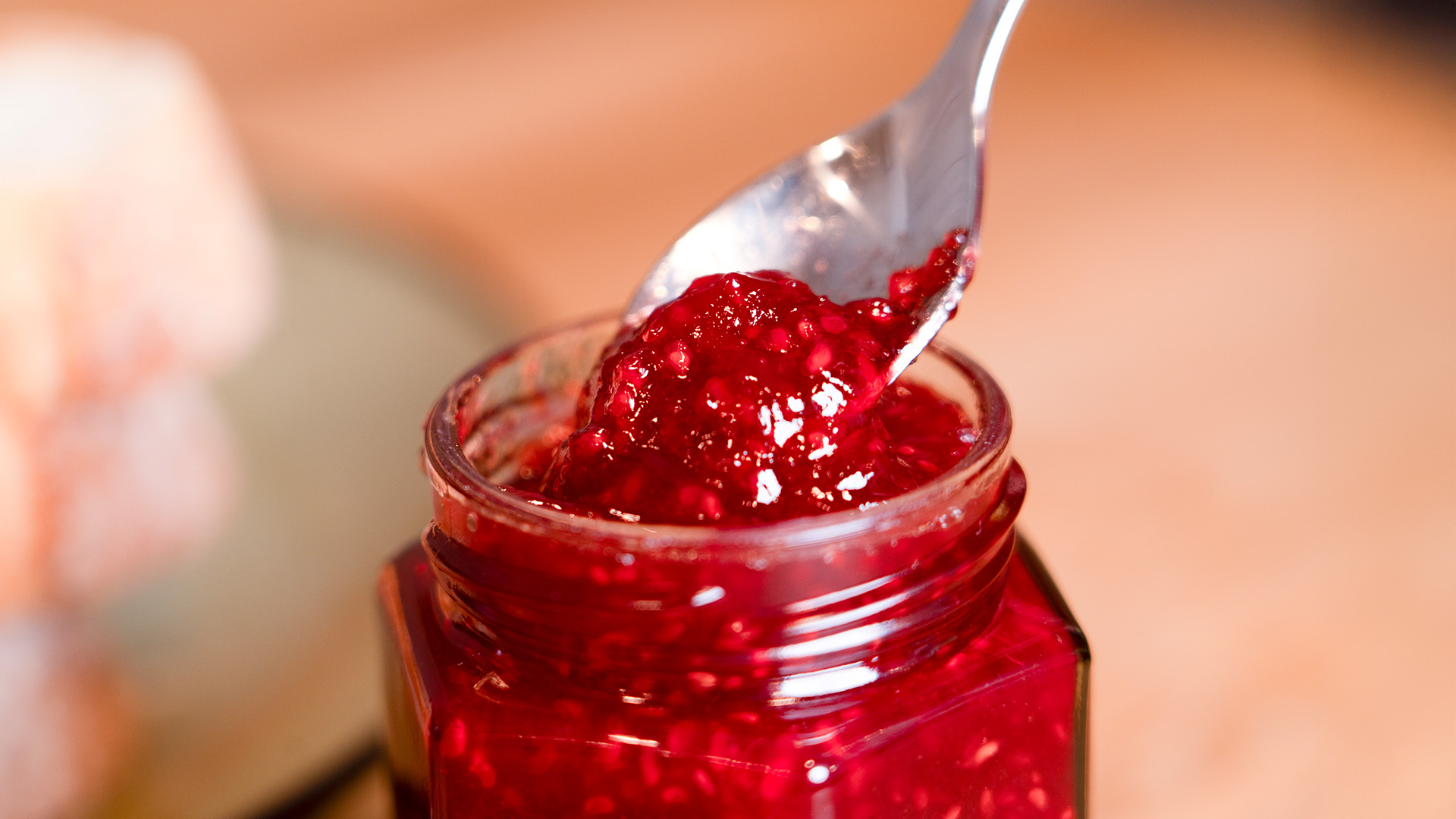 Großaufnahme eines Marmeladenglases ohne Deckel, aus dem ein Löffel frische Konfitüre entnommen wird.