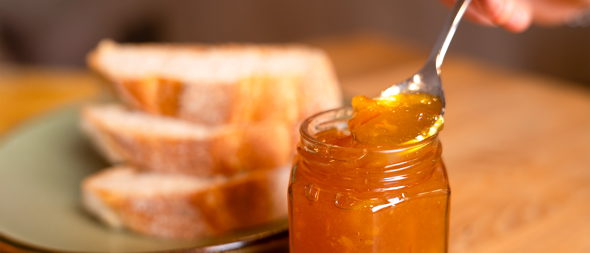 Glas Schwarzwälder Konfitüre, aus dem ein Löffel frische Marmelade entnommen wird; im Hintergrund ein Teller mit drei Brotscheiben.
